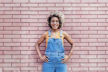 Gülümseyen siyah kadın elleri belinde dışarıda tuğladan duvara karşı duruyor.