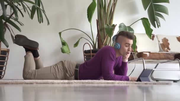 Giovane ragazzo carino moderno utilizzando tablet con cuffie per avere una conversazione videocall online con amici o familiari sul pavimento a casa — Video Stock