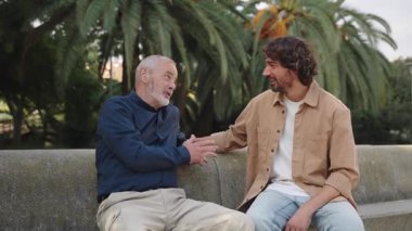 Adult son supporting and listening senior father while talking sharing good news sitting together in a park 