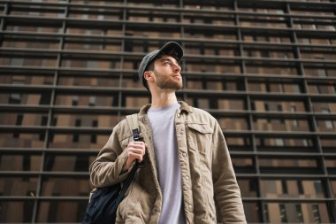 Portrait young man with backpack and cap standing in the city looking to the side with confident expression