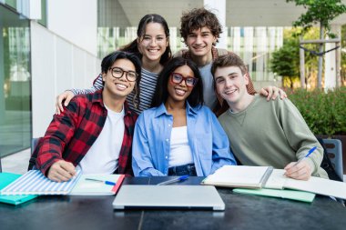 Neşeli uluslararası değişim üniversitesi öğrencileri kampüs kütüphanesinde çalışırken kameraya bakıp gülümsüyor.