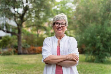 Şehir parkında kollarını kavuşturmuş gülümseyen son sınıf öğrencisi kadın.