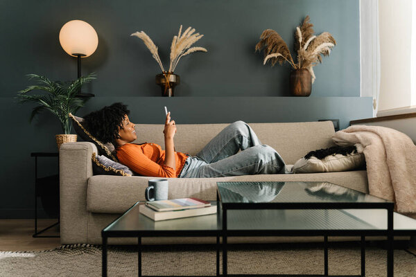 Relaxed African American woman lying on a sofa at home using mobile phone app and social media