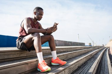 Eğitimden sonra oturan Afro-Amerikalı adam, akıllı kasetteki fitness seviyelerini kontrol ediyor ve cep telefonu uygulaması kullanıyor.