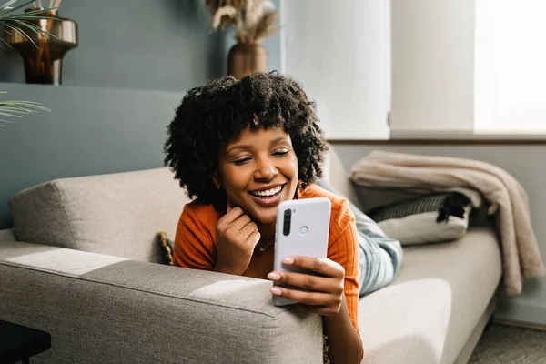 Sosyal medyada içeriği paylaşmak için cep telefonu uygulamasını kullanarak evdeki koltukta dinlenen gülümseyen Afrikalı Amerikalı kadın.