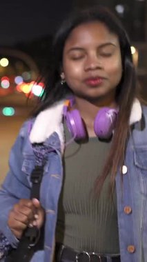 Geceleri şehirde dolaşan, sırt çantası ve kulaklık taşıyan, kendinden emin, Afro-Amerikan genç bir kadın.