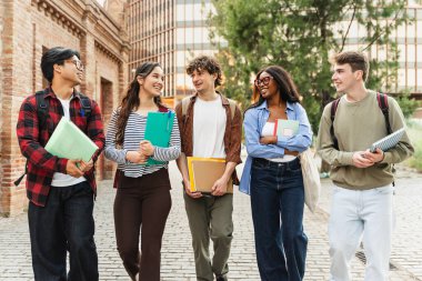 Çok kültürlü üniversite öğrencileri ellerinde not defterleriyle birlikte yürüyorlar. Açık hava kampüs yaşam tarzı.