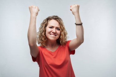 Kıvırcık saçlı, sıradan bir tişört giymiş, gülümseyen ve kazanan bir jest yapan, harika haberleri kutlayan mutlu bir kadın resmi. Stüdyo çekimi, beyaz arkaplan, izole edilmiş