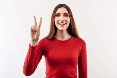 Uzun kestane rengi saçları olan mutlu genç bir kadının portresi barış jesti gösteriyor, günlük kırmızı süveter giyiyor. Stüdyo fotoğrafı, beyaz arka plan. İnsani duygular, yüz ifadesi kavramı