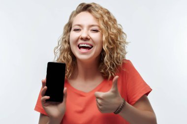 Kıvırcık saçlı, günlük tişörtlü, Akıllı Telefon Boş Ekran Gösteren, Kamera Tavsiye Eden Mobil Uygulama 'ya bakan genç bir kadın.