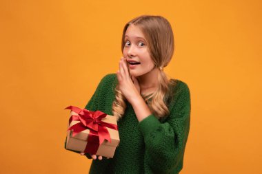 12-14 yaşlarında, sıcak yeşil kazaklı, elinde kırmızı fiyonklu hediye kutusu olan çekici bir sarışın kız resmi. Stüdyo fotoğrafı, sarı arka plan, izole edilmiş. Yeni yıl kadınlarının doğum günü konsepti