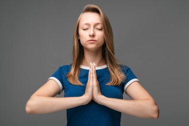 Genç sarışın kadın yoga yapıyor namaste 'de el ele tutuşuyor ve gözlerini kapalı tutuyor. Evde meditasyon yapıyor, huzur ve sevgi için dua ediyor, sakin ve huzurlu bir yüz ifadesi var.