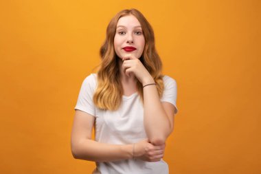Dalgalı kızıl saçlı, eli çenesinde duran ve sarı arka planda izole edilmiş bir kameraya dikkatlice bakan dalgın genç bir kadının fotoğrafı. Düşünce kavramı