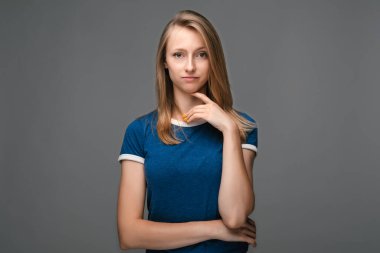 Sarışın, düz saçlı ve mavi tişörtlü düşünceli genç bir kadın. Gri arka planda izole edilmiş, el kaldırılmış bir şekilde kameraya bakıyor. Düşünce kavramı