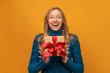 Elinde kurdeleyle süslenmiş bir hediye tutan mutlu genç bir kadının portresi. Stüdyo çekimi, sarı arkaplan