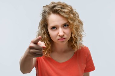 Sıradan tişörtlü, kıvırcık saçlı güzel genç bir kadın kameraya bakıyor ve parmağını sana doğrultuyor. Stüdyo fotoğrafı, beyaz arka plan. İnsani duygular, yüz ifadesi kavramı