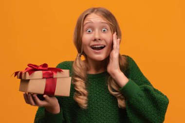12-14 yaşlarında, sıcak yeşil süveter giyen, gülümseyen ve kırmızı kurdeleli hediye kutusunu tutan çekici bir sarışın kız resmi. Stüdyo fotoğrafı, sarı arka plan. Yeni Yıl Kadınların Doğum Günü Tatil Konsepti