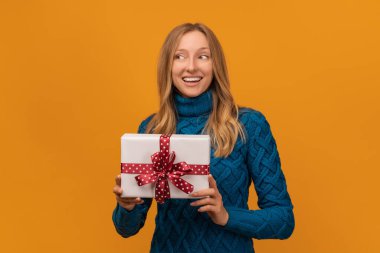 Sıcak örgü süveter giymiş, gülümseyen ve elinde kırmızı kurdeleyle hediyesini tutan alımlı genç bir kadın resmi. Stüdyo fotoğrafı, sarı arka plan. Yeni Yıl, Kadınlar Günü, Doğum günü, Tatil konsepti