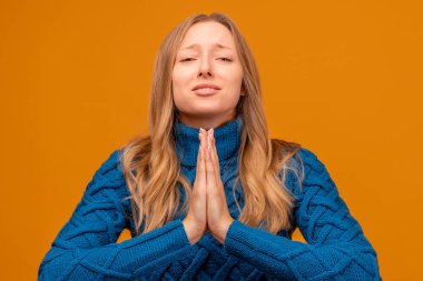 Mavi kazaklı genç bir kadının portresi avuç içlerini birleştirip dua ediyor. Meditasyon yapıyor, huzur için dua ediyor sakin ve huzurlu bir yüz ifadesi var. Stüdyo çekimi, sarı arkaplan