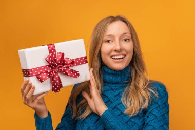 Sıcak örgü süveter giymiş, gülümseyen ve elinde kırmızı kurdeleyle hediyesini tutan alımlı genç bir kadın resmi. Stüdyo fotoğrafı, sarı arka plan. Yeni Yıl, Kadınlar Günü, Doğum günü, Tatil konsepti