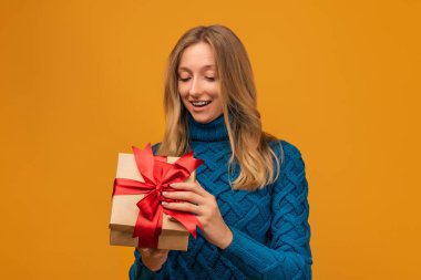 Çekici, mavi kazaklı sarışın kadın kırmızı kurdeleli hediyesini açıyor. Stüdyo fotoğrafı, sarı arka plan. Yeni Yıl, Kadınlar Günü, Doğum günü, Tatil konsepti