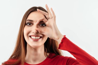 20 'li yaşlardaki güzel bir kadının parmaklarının açtığı delikten sana bakıp güldüğünü hayal et. Stüdyo fotoğrafı, beyaz arka plan. İnsani duygular, yüz ifadesi kavramı