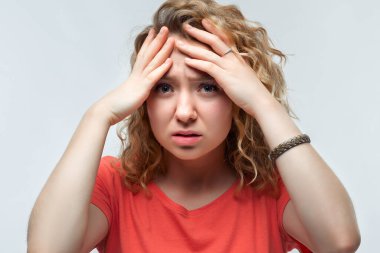 Kafasını kavrayan ve baş ağrısı yüzünden şakaklarını ovuşturan sıradan tişörtlü, kıvırcık saçlı üzgün bir kadının fotoğrafı. Stüdyo fotoğrafı, beyaz arka plan. İnsani duygular, yüz ifadesi kavramı