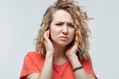 Kıvırcık saçlı, sıradan tişörtlü, kulaklarını parmaklarıyla tıkayan korkunç sesler duyan, rahatsızlık veren sarışın kadın portresi. İnsan duyguları konsepti. Stüdyo çekimi, beyaz arkaplan