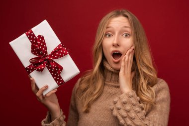 Sıcak süveter giyen, gülümseyen ve elinde kırmızı kurdeleyle hediyesini tutan alımlı sarışın bir kadın resmi. Stüdyo kırmızı zemin çekti. Yeni yıl kadınlarının doğum günü konsepti
