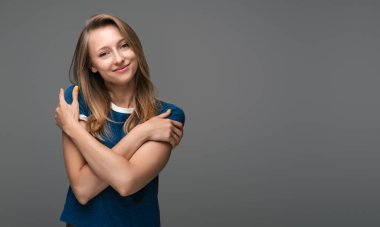 Charming blonde girl hugging herself hold shoulders, tilting head, smiling flirty romantic attitude, standing delighted gray background. Love, care tenderness concept. Copy space