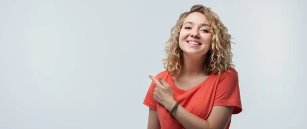 巻き毛の若い幸せな女性のイメージは カジュアルなTシャツを着て 笑顔と額を脇に指して 広告のための場所を示しています スタジオショット 白い背景 隔離された — ストック写真