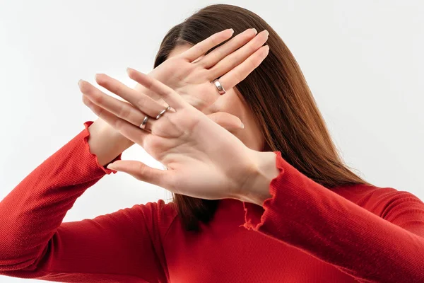 Image of serious woman with long chestnut hair in casual red sweater gestures actively hands at camera, stop gesture with hands. No way, stop doing. Studio shot, white background. Human emotions concept