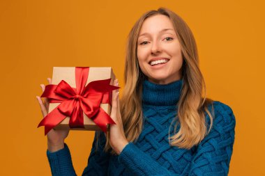 Sıcak örgü süveter giymiş, gülümseyen ve elinde kırmızı kurdeleyle hediyesini tutan alımlı genç bir kadın resmi. Stüdyo fotoğrafı, sarı arka plan. Yeni Yıl, Kadınlar Günü, Doğum günü, Tatil konsepti