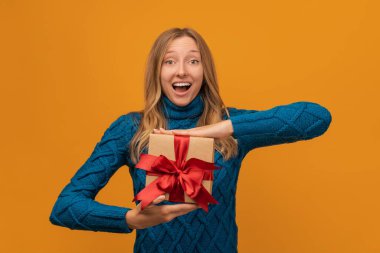 Sıcak örgü süveter giymiş, gülümseyen ve elinde kırmızı kurdeleyle hediyesini tutan alımlı genç bir kadın resmi. Stüdyo fotoğrafı, sarı arka plan. Yeni Yıl, Kadınlar Günü, Doğum günü, Tatil konsepti