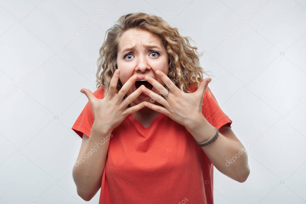 I'm afraid. Image of scared blonde girl with curly hair in casual t shirt covering her mouth with hands. Fright, phobia, panic attack, horror and facial expression concept