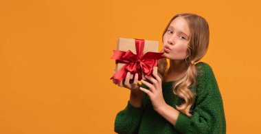 12-14 yaşlarında, sıcak yeşil kazaklı, elinde kırmızı fiyonklu hediye kutusu olan çekici bir sarışın kız resmi. Stüdyo fotoğrafı, sarı arka plan, izole edilmiş. Yeni yıl kadınlarının doğum günü konsepti