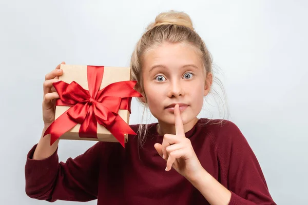 12-15 yaşlarında sıradan bir süveter giymiş, elinde kurdeleyle süslenmiş bir hediye taşıyan mutlu sarışın kızın portresi. Stüdyo beyaz arka plan çekti, izole. Yeni Yıl, Kadınlar Günü, Doğum günü, Tatil konsepti
