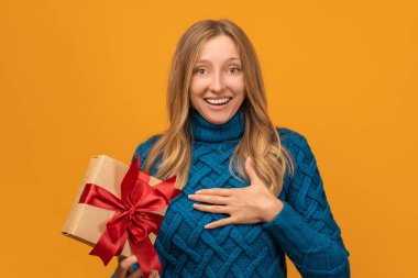 Sıcak örgü süveter giymiş, gülümseyen ve elinde kırmızı kurdeleyle hediyesini tutan alımlı genç bir kadın resmi. Stüdyo fotoğrafı, sarı arka plan. Yeni Yıl, Kadınlar Günü, Doğum günü, Tatil konsepti