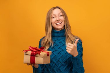 Çekici, mavi kazaklı sarışın kadın kırmızı kurdeleli hediyesini açıyor. Stüdyo fotoğrafı, sarı arka plan. Yeni Yıl, Kadınlar Günü, Doğum günü, Tatil konsepti