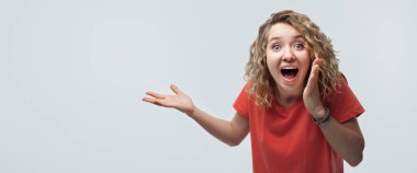 Şok olmuş, kıvırcık saçlı, sıradan tişörtlü, kameraya sürpriz yapan genç bir kadın. İnsani duygular, yüz ifadesi kavramı. Stüdyo çekimi, beyaz arkaplan