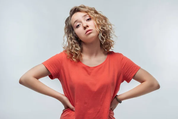Kaşlarını çatan ve kalçalarında el ele tutuşan, sıradan tişörtlü, kıvırcık saçlı, sinirli sarışın kızın portresi. Stüdyo fotoğrafı, beyaz arka plan. İnsani duygular, yüz ifadesi kavramı
