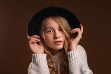 Siyah şapkalı çekici sarışın kız stüdyoda kahverengi arka planda poz veriyor. Güzellik ve zarafet kavramı
