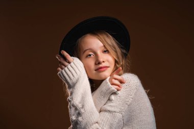 Siyah şapkalı çekici sarışın kız stüdyoda kahverengi arka planda poz veriyor. Güzellik ve zarafet kavramı