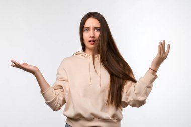 Uzun kestane rengi saçları olan, bej kapüşonlu şık giyinen, şüpheyle el ele tutuşan güvensiz genç bir kadının stüdyo fotoğrafı. Şüphe, belirsizlik ve karmaşa kavramı