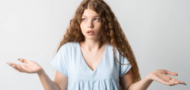 Ne oldu? Kafası karışmış, uzun kıvırcık saçlı güzel bir kızın portresi, ellerini yana açıyor, sırıtıyor, seçimler yaparken şüpheye düşüyor, gündelik tişört giyiyor. Beyaz arkaplanda izole.