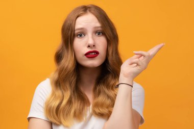Studio shot of displeased girl with wavy redhead, wearing white t-shirt, gesturing with raised palm, frowning, being displeased and confused with dumb question. Human emotions concept