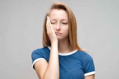 Yorgun, sarı saçlı, yüzünü kapatan, yorgunluk çeken, iyice dinlenmesi gereken, mavi tişörtlü, uykulu bir yüzü olan, gri arkaplan üzerinde izole edilmiş genç bir kadının fotoğrafı. Yorgunluk ve insanlar