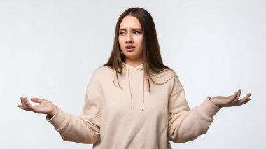 Uzun kestane rengi saçları olan, bej kapüşonlu şık giyinen, şüpheyle el ele tutuşan güvensiz genç bir kadının stüdyo fotoğrafı. Şüphe, belirsizlik ve karmaşa kavramı