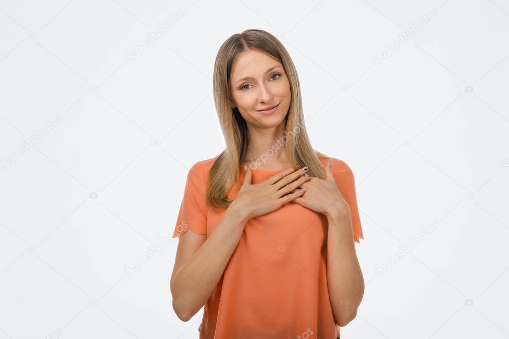 Pleasant young blond woman in casual clothes hold hands on heart cheerfully thanking for compliments or praises, feel grateful and pleased. Studio shot, white background