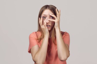 Endişeli ve endişeli, sıradan giysili sarışın kız korkmuş bir ifadeyle bakıyor. Stüdyo çekimi, gri arkaplan